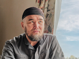 Portrait of senior asian muslim man with beard wearing skullcap sitting by window.
