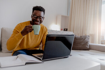 African young man in eyeglasses sitting at the table in front of laptop computer and looking at monitor at home, drinking tea. Handsome young African man using laptop and smiling while sitting indoors