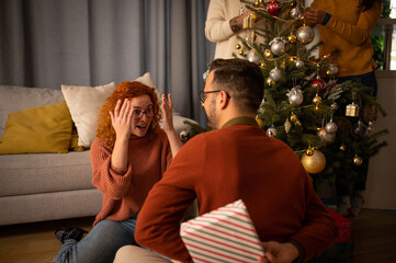 Young couple in love sitting on a sofa next to a Christmas tree. The guy gives the woman a gift for the new year