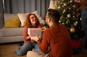 Obraz premium Young couple in love sitting on a sofa next to a Christmas tree. The guy gives the woman a gift for the new year