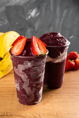 two Brazilian Frozen Açai Berry Ice Cream Smoothie in plastic cup with Strawberries and condensed milk. On a wooden desk and a gray summer background with fruits. Front view for menu and social media