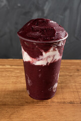 Brazilian Frozen Açai Berry Ice Cream Smoothie in plastic cup with Condensed Milk. On a wooden desk and a gray summer background. Front view for menu and social media