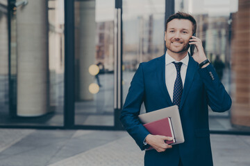 Smiling male company worker leaving office building while talking on phone with his colleague