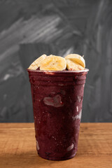 Brazilian Frozen Açai Berry Ice Cream Smoothie in plastic cup with Bananas. On a wooden desk and a gray summer background. Front view for menu and social media