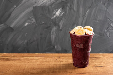 Brazilian Frozen Açai Berry Ice Cream Smoothie in plastic cup with Bananas. On a wooden desk and a gray summer background. Front view for menu and social media
