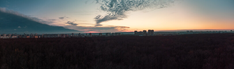 Aerial sunset evening wide panorama view above dark blue autumn forest near residential Pavlove Pole district in Kharkiv city. Gray multistory buildings with cloudy sky on horizon