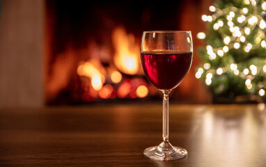 Red wine glass, burning fireplace background. Christmas tree bokeh lights decoration