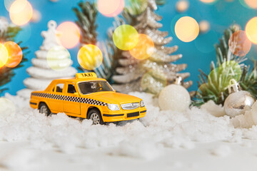 Christmas banner Background. Yellow toy car Taxi Cab model and winter decorations ornaments on blue background with snow and defocused garland lights. City traffic delivery taxi service concept
