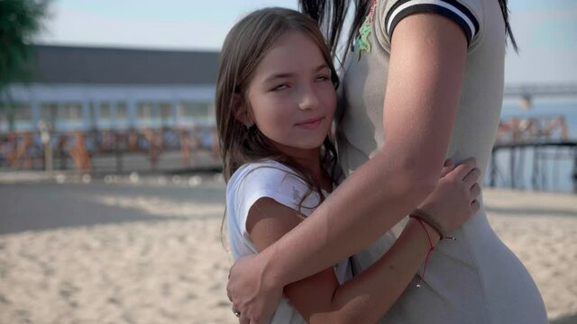 Portrait of happy mother and daughter 9 years standing hugging against the backdrop near beach in sunlight and smiling at camera enjoying moment