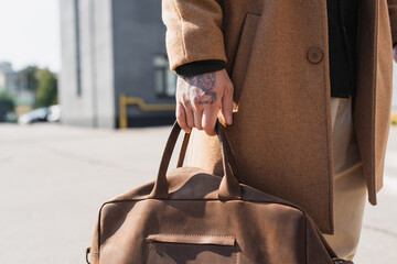 partial view of tattooed man in beige coat holding leather briefcase outdoors
