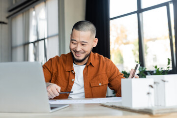smiling asian architect holding smartphone while working with blueprints near blurred laptop - obrazy, fototapety, plakaty