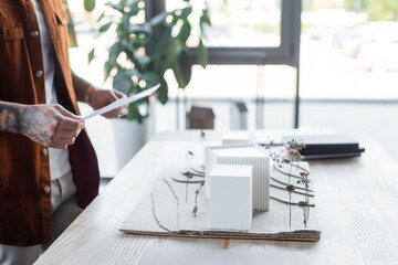 partial view of tattooed engineer holding document near blurred house models on desk