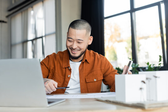 Smiling Asian Architect Holding Smartphone While Working With Blueprints Near Blurred Laptop