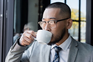 asian businessman in formal wear and eyeglasses drinking coffee in office
