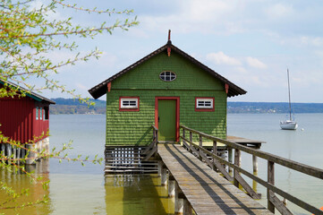 a long wooden pier leading to colorful boat houses and a sailing boat in background on sunlit and serene lake Ammersee in German fishing village Schondorf on sunny spring day (Bavaria, Germany)	