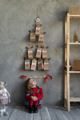 Happy girl looking at advent calendar on the wall sitting on the floor in the room, handmade christmas decor, horizontal view, kid festive fan.