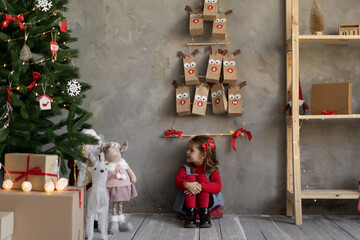 Fototapeta na wymiar Little girl with a Christmas tree in the living room with an advent calendar hanging on the wall in craft bags, gifts and festive decor on the eve.