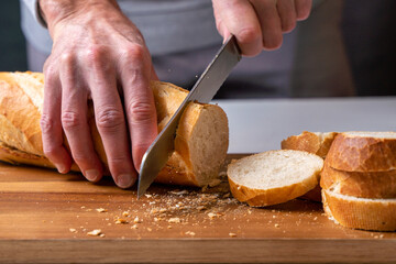hands cutting a freshly baked baguette into slices on a wooden board, front view