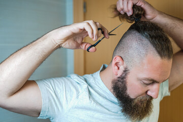 Obraz na płótnie Canvas Caucasian man with long hair and beard cuts his ponytail with scissors.