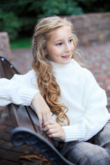 Obraz premium Blond girl on a bench in the park. Portrait of a blue-eyed girl in a white sweater in the park.