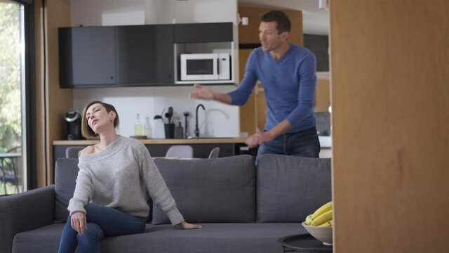 Slim Woman Sitting On Couch Standing Up Walking Away As Aggressive Angry Man Shouting Gesturing. Portrait Of Beautiful Caucasian Wife Leaving Furious Husband In Living Room Alone