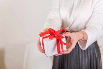 Female hands holding small gift with ribbon.Girl in white shirt holding a white gift box with a red bow on blurred background. Holidays concept.Banner for Christmas, hew year, birthday concept.