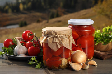 Glass jars with pickled tomatoes and fresh ingredients on wooden table in nature