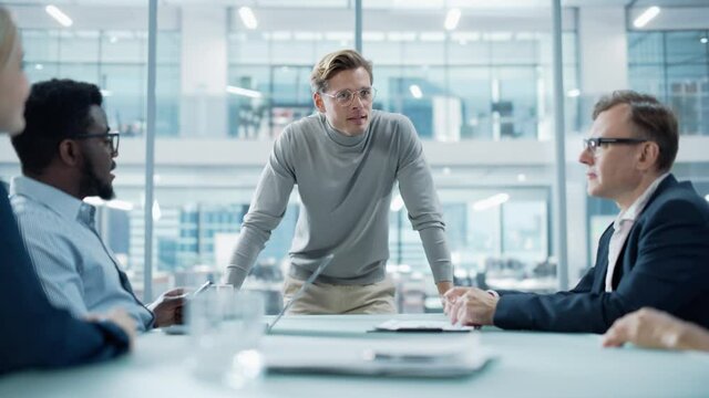 Modern Office Meeting Room: Handsome Executive Director Stands in the Head of the Conference Table Leans on it and Delivers Eloquent Speech to a Group of Businesspeople and Investors.