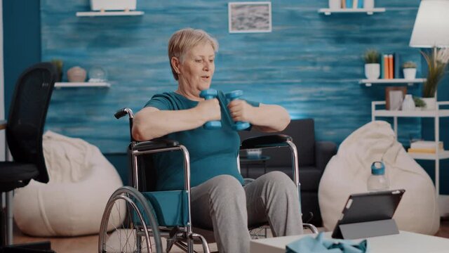 Senior adult sitting in wheelchair and doing exercise with weights while following training video on tablet. Elder woman with disability using dumbbells to train with online lesson.