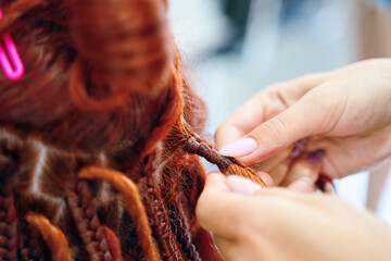 Close up of braiding process african plaits with colored kanekalon. Hairdresser weaves woman ginger dreadlocks. Bright pigtails on head. Hippie style hairstyle. Beauty salon services.