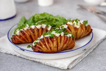 Bacon Stuffed Hasselback Potatoes topped with creamy sauce and fresh chive. Portion served on white plate. Close up, selective focus, gray concrete background.