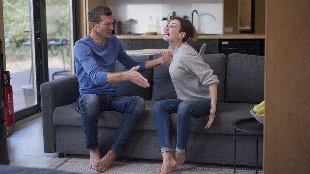 Wide shot angry couple shouting gesturing sitting on couch in living room at home. Caucasian man and woman yelling arguing indoors screaming. Imbalance and conflicts concept