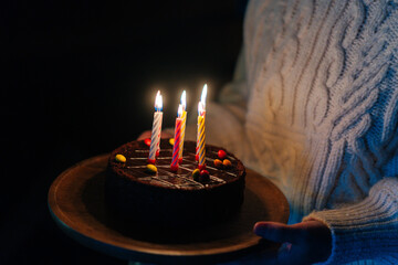 Close-up cropped shot of unrecognizable young woman holding in hands birthday cake with six burning candles in dark room. Female preparing to wish happy birthday to child, husband or friend.