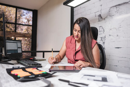 Hermosa mujer de negocios latina sentada en su oficina con un lápiz en su mano tomando apuntes de sus proyectos de arquitectura