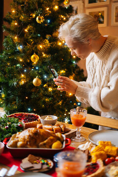 Vertical shot of attractive young woman taking photo or making video of beautiful Christmas dinner table on smartphone, on blurred background of decorating xmas tree and celebration bokeh lights.