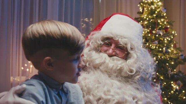 Close up of little cute boy sitting on Santa's knees making a wish. Whitebearded Santa is hugging him and asking him questions.