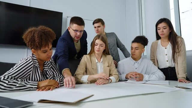 Team of designers discussing masterplan lying on table, some people sitting, some standing behind. Tracking shot multiethnic group having discussion. Concept of business