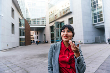 Smiling Hispanic businesswoman sending voice messages through her mobile phone near a business and office building.