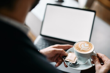 Close-up of businessman enjoys in cup of coffee while using laptop in cafe.