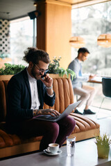 Happy entrepreneur works on laptop while making phone call in cafe.
