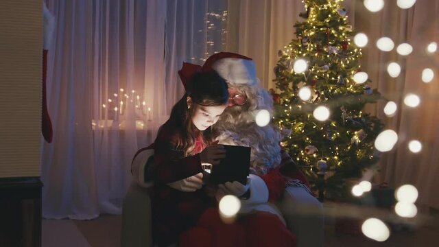 Little cute girl sitting on Santa's knees, opening a gift in front of Christmas tree. Adorable child is happy and surprised, hugging Santa Claus.
