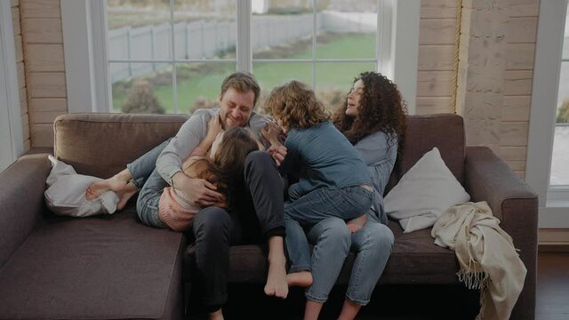 Young family of 4 sit together on a couch tickling and laughing, wearing denim. Family spends time together. Happy family sits at home in lockdown. 50 fps. High quality 4k footage