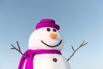 Funny snowman in stylish brown hat and pink scalf on snowy field. Blue sky on background