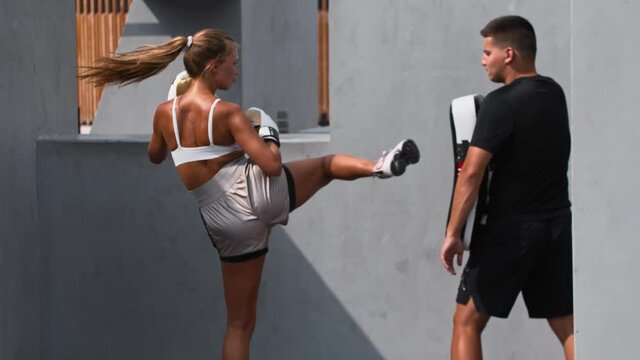 Young woman having a boxing training with her coach outside - kicking the portative punching bag 