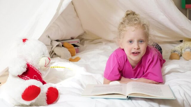 a cute girl with curly hair reads a book in a hut from a sheet on the bed, little girl having fun and playing in her tent, Hut in the kids room, activities concept