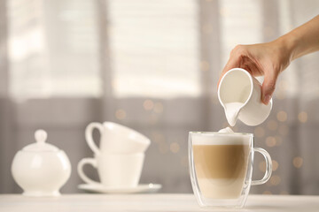 Woman pouring milk into cup with coffee at table indoors, closeup