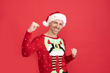 Happy guy in Santa Claus hat and Christmas jumper make winning gesture red background, new year