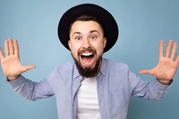 Close-up photo shot of positive funny joyful happy smiling good looking young brunet bearded man wearing casual blue shirt and white t-shirt and stylish black hat poising isolated on blue background