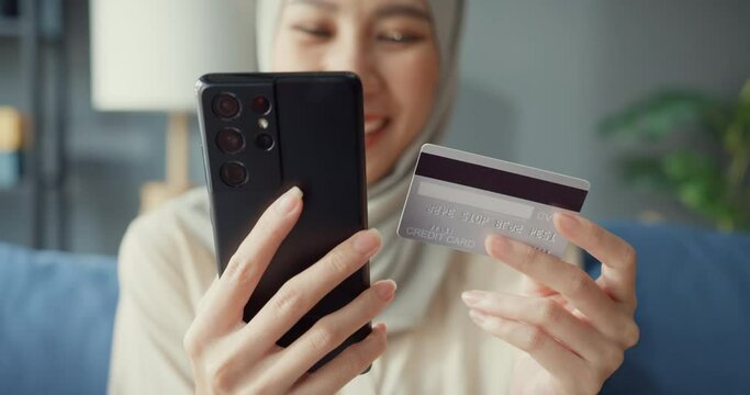 Close-up Attractive cheerful young Asian islam muslim beauty girl in hijab with casual sitting on couch use phone enjoy successful payment online shopping with credit card in cozy living room at home.