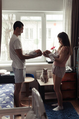 Young family with newborn twins. Yong parents standing face to face near the window. Cute and tender pfoto of twins sleeping in mother's and father's arms. Siluette photo. Selective focus. Film grain